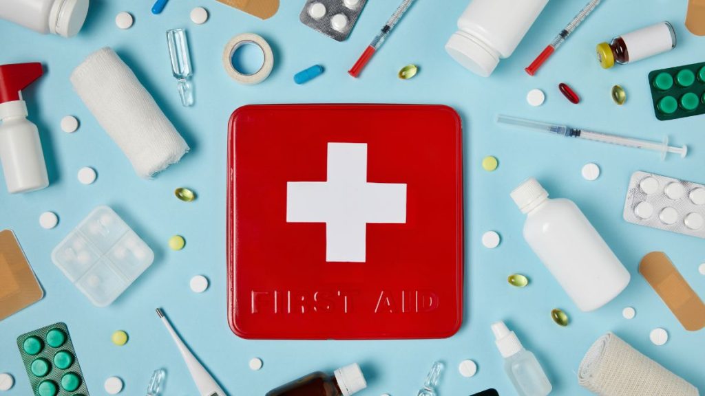 a red first aid box in the centre with pills, bandages, medical tape and more surrounding the box on a light blue background.