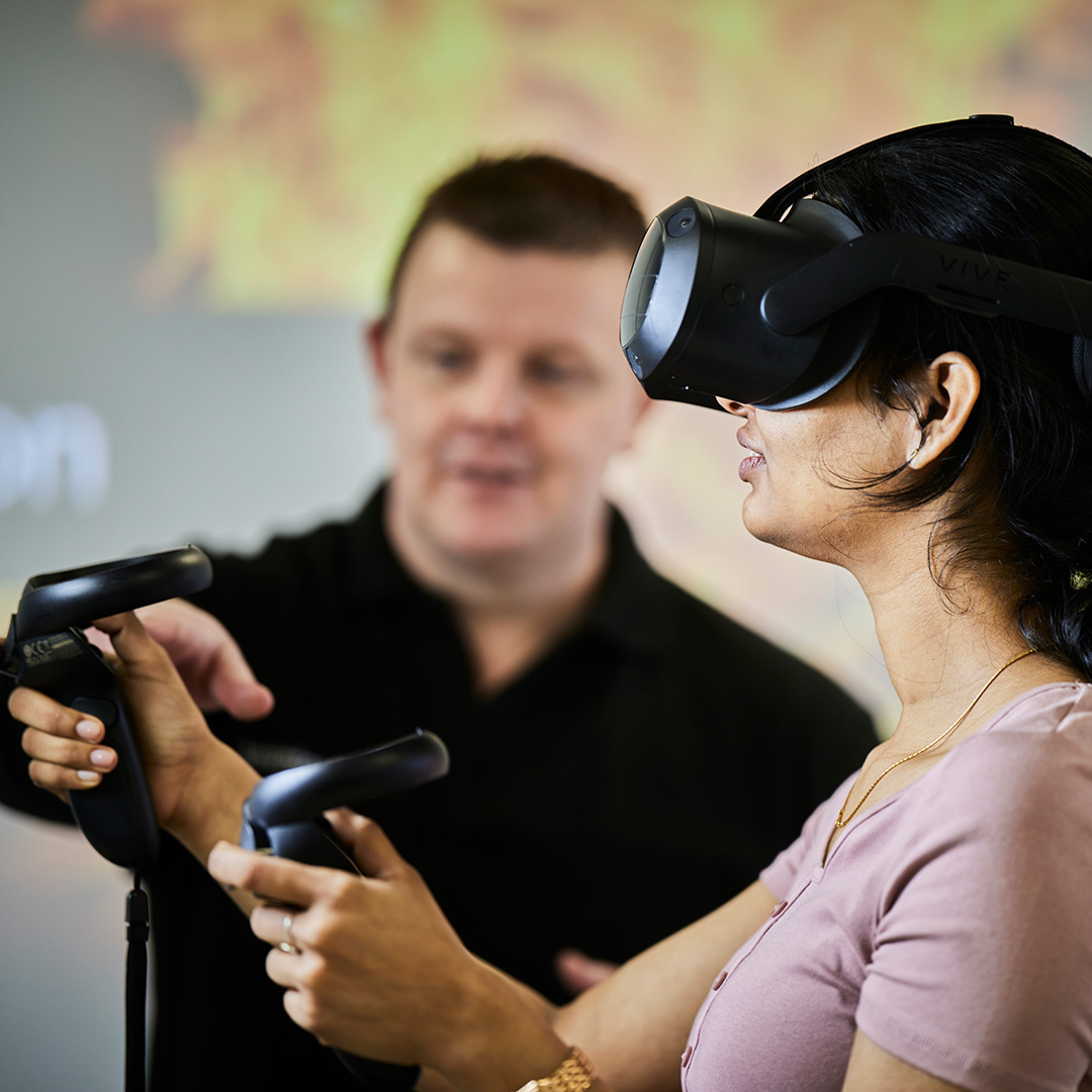 A young adult women with tied up black hair using a black VR headset and two black VR controllers with a fire safety training instructor blurred in the background pointing to one of the black VR controllers.