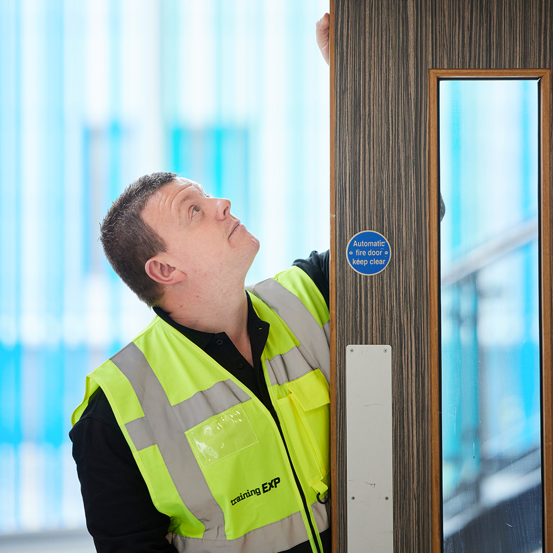 An adult man looking up towards his hand at the top of a fire door.