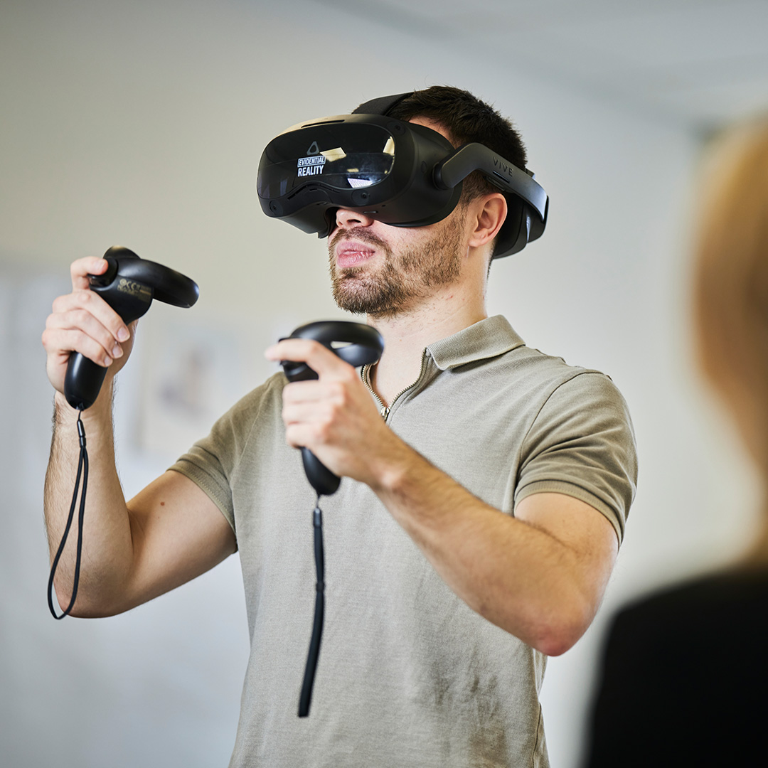 A young adult male using a black VR headset and two black VR controllers in a classroom training environment.