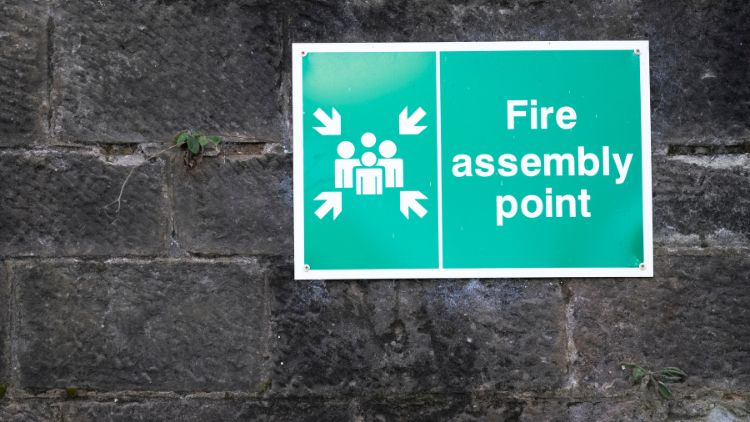 An image of a grey brick wall with a green sign on it reading 'Fire Assembly Point'.